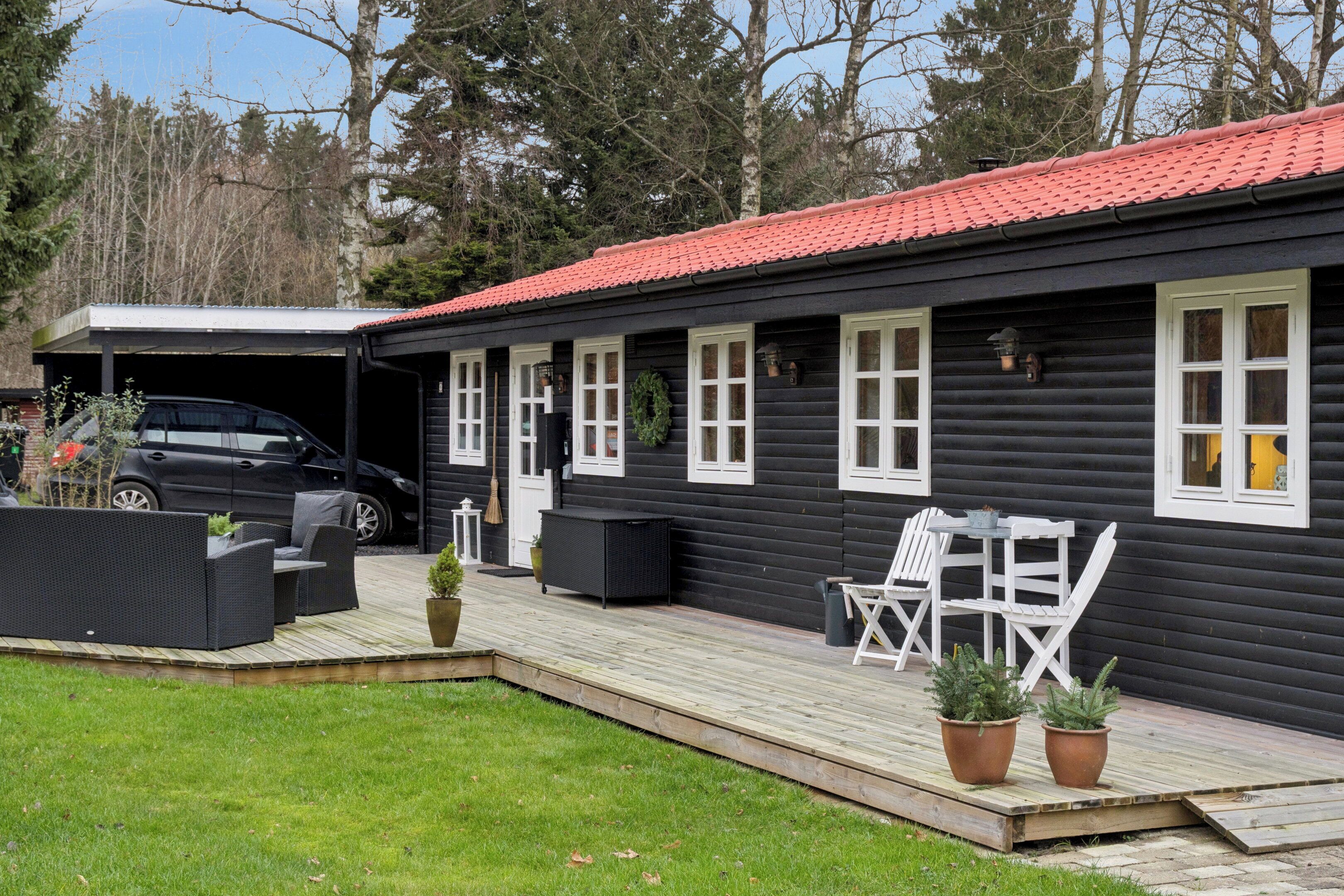 A black wooden house with a red roof, a covered carport, outdoor furniture, and potted plants.