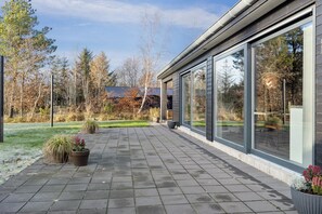 A paved patio with potted plants, large windows, and a view of trees and a building in the background. - Lovely holiday home near Hals in North Jutland (Hals)