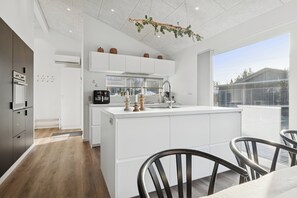 A modern kitchen with white cabinetry, a central island, and a dining area with metal chairs. - Lovely holiday home near Hals in North Jutland (Hals)