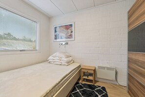Een kleine eenpersoonskamer met raam, radiator, een lijst met een afbeelding en een houten kastje.