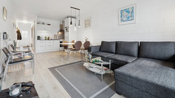 Un salon moderne avec un canapé gris, un coin repas avec une table et des chaises, et une cuisine en arrière-plan.