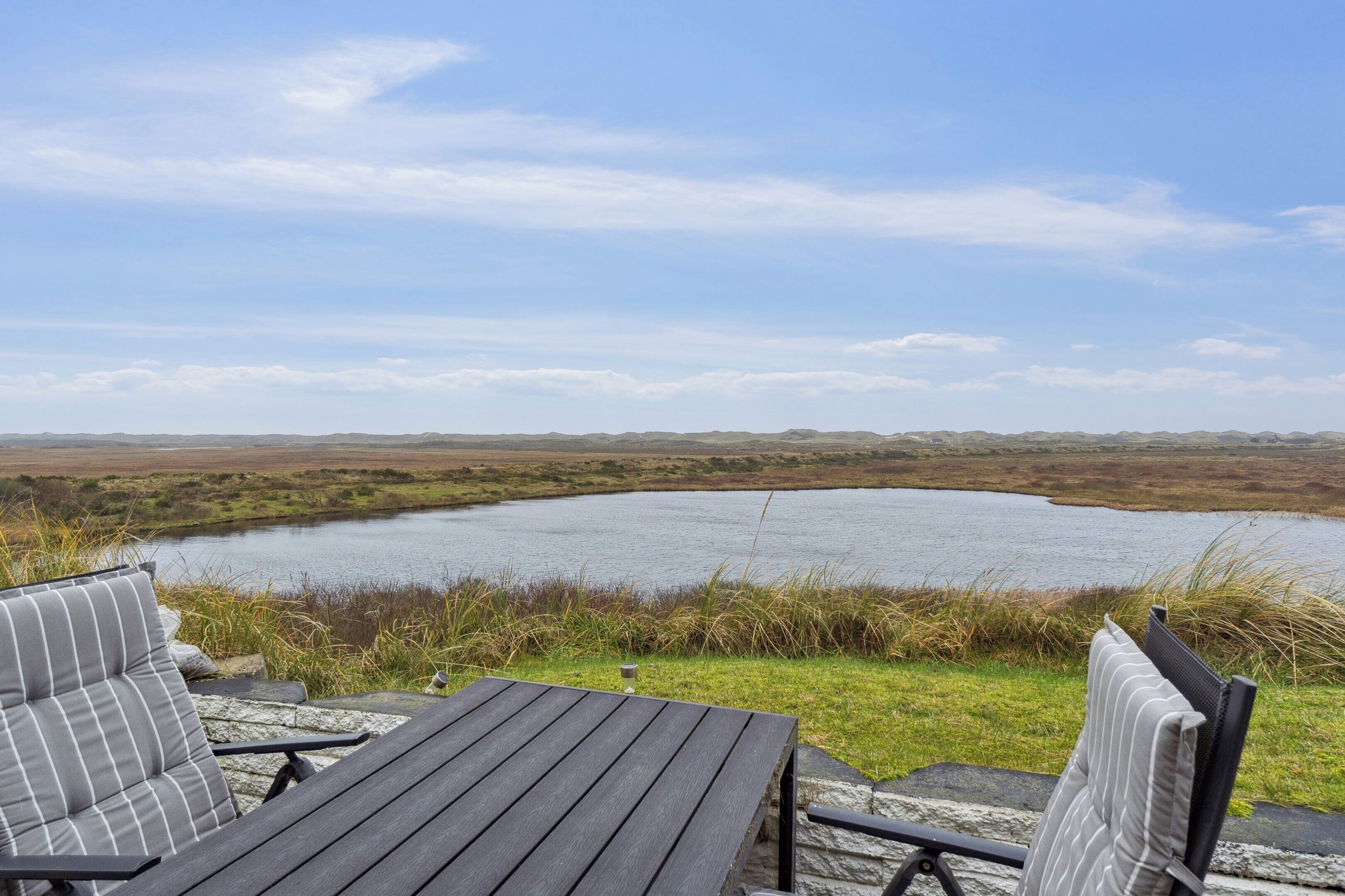 Una mesa de madera y dos sillas a rayas frente a un lago, con un paisaje cubierto de hierba al fondo.