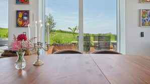 Un comedor con una mesa, sillas, un jarrón con flores y vistas a un paisaje verde a través de amplias ventanas.
