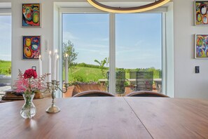 Ein Essbereich mit Tisch, Stühlen, einer Blumenvase und Blick auf eine grüne Landschaft durch große Fenster.