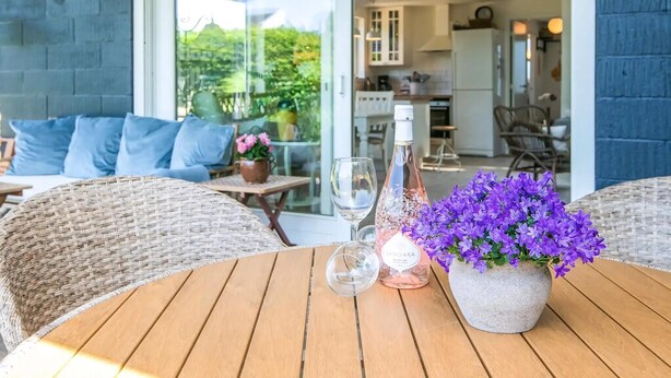 Ein Holztisch mit einer Vase voller lila Blumen, einer Weinflasche und einem Glas auf einer Terrasse mit Korbmöbeln und Blick auf die Küche.