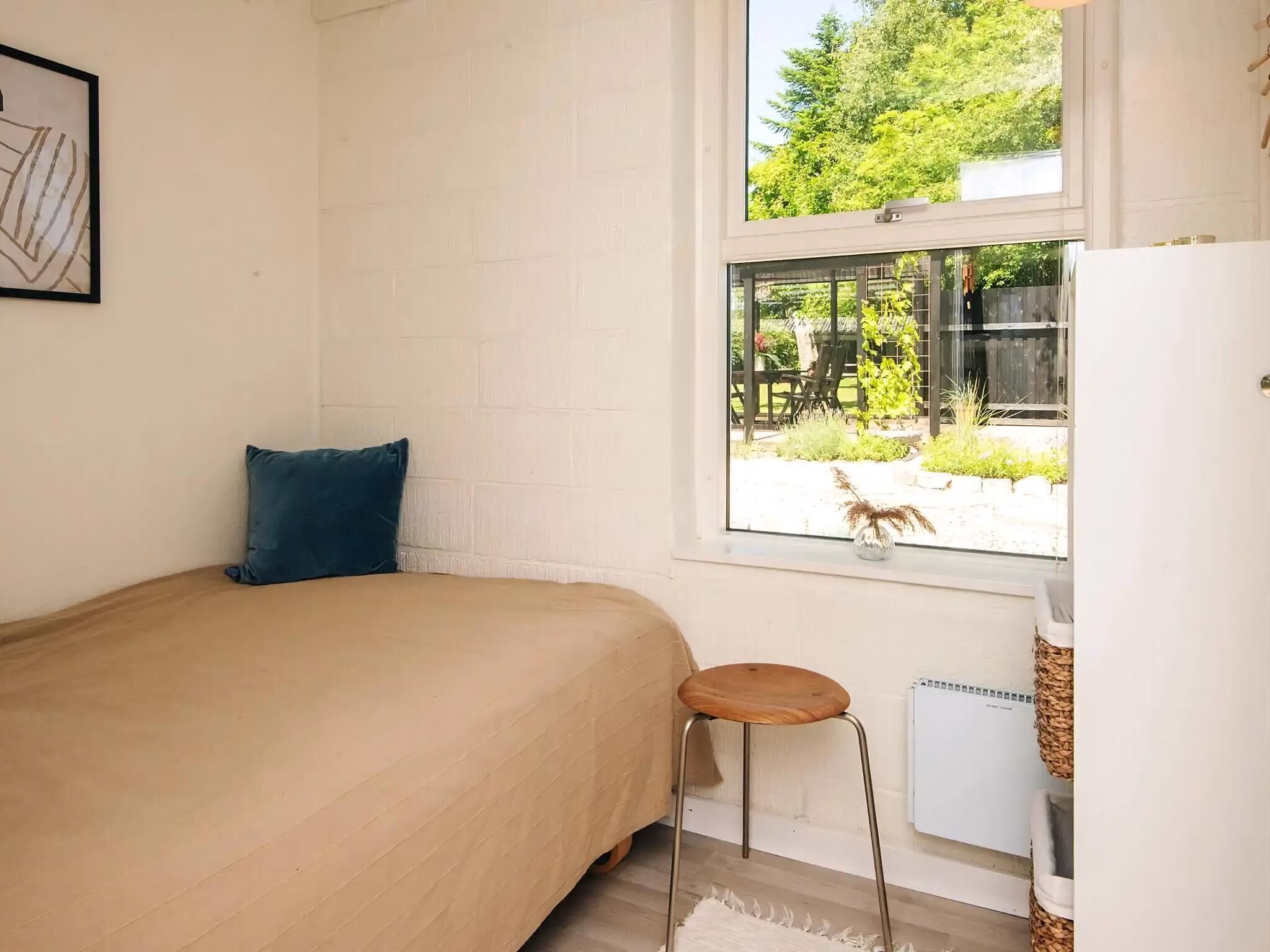 Een kleine, lichte kamer met een bed, een krukje, een raam en een radiator.