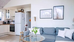 Ein modernes Wohnzimmer mit einem grauen Sofa, einem runden Glastisch mit gestreiften Vasen und einem Korbstuhl. Im Hintergrund ist eine Küche mit weißen Schränken und einem Kühlschrank zu sehen.