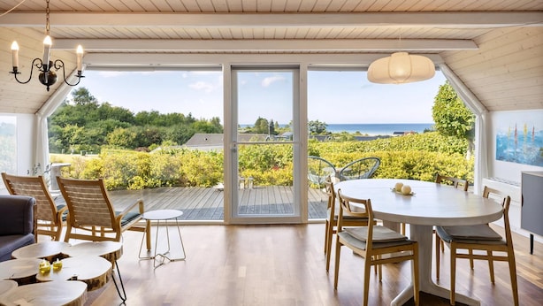 A spacious room with a large sliding glass door leading to a deck with a scenic view. Inside, there is a round dining table with chairs, a sofa, and a chandelier. - Idyllic summer house with sea view (Glesborg)
