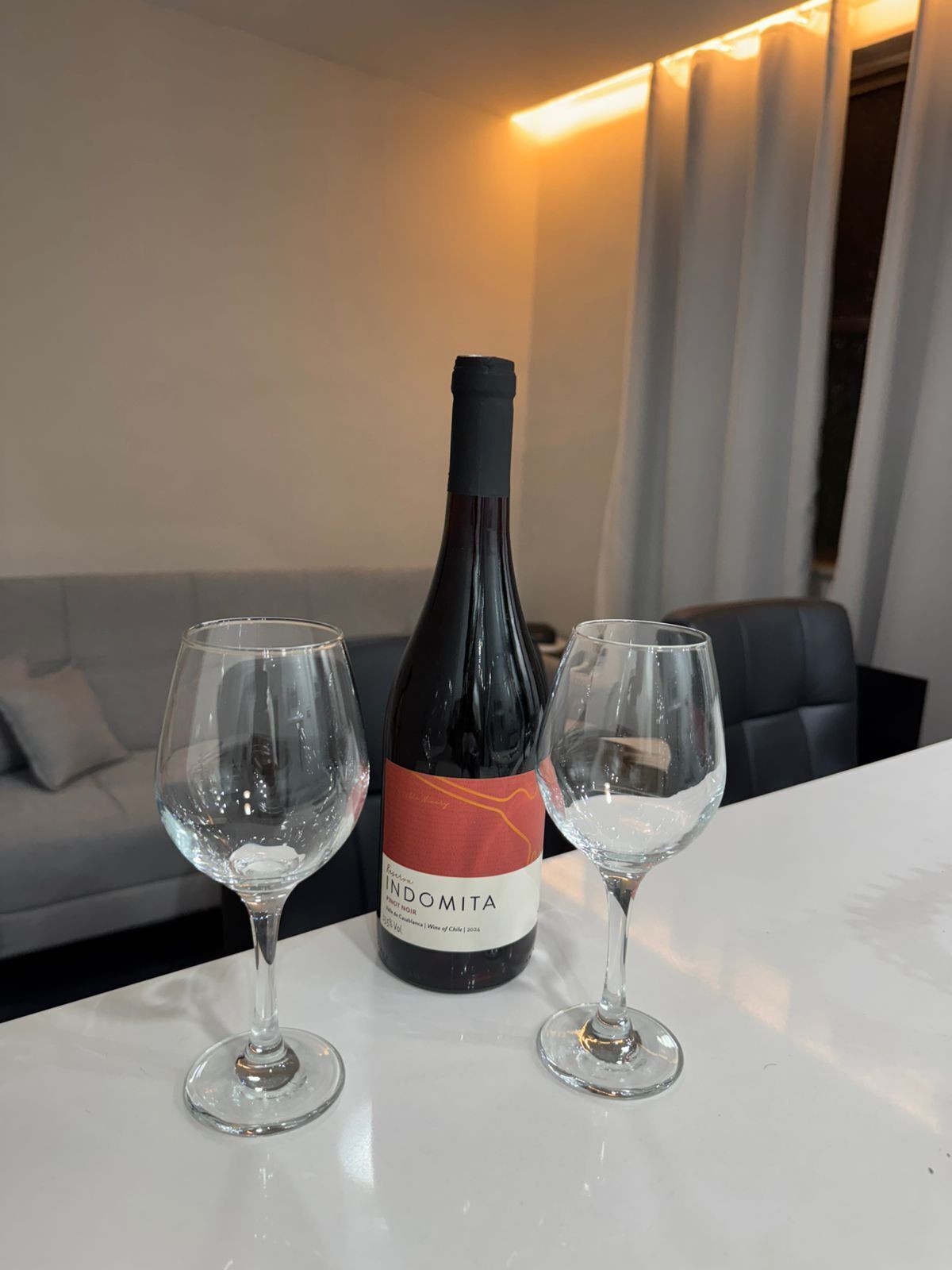 Una botella de vino y dos copas sobre una mesa en una habitación con sofá y cortinas.