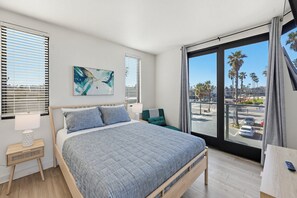 A bedroom with a bed, bedside tables, a nightstand, a window with blinds, a view of palm trees and a parking lot. - Ocean Park Combo 9 & 10 (San Diego)