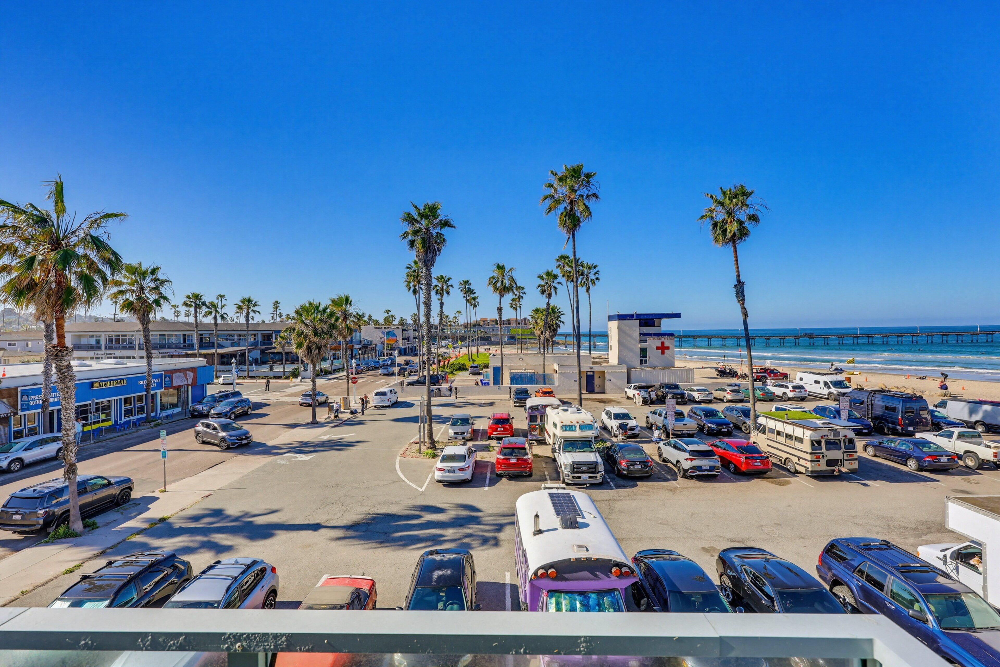 Un estacionamiento con varios vehículos, palmeras y una playa al fondo.