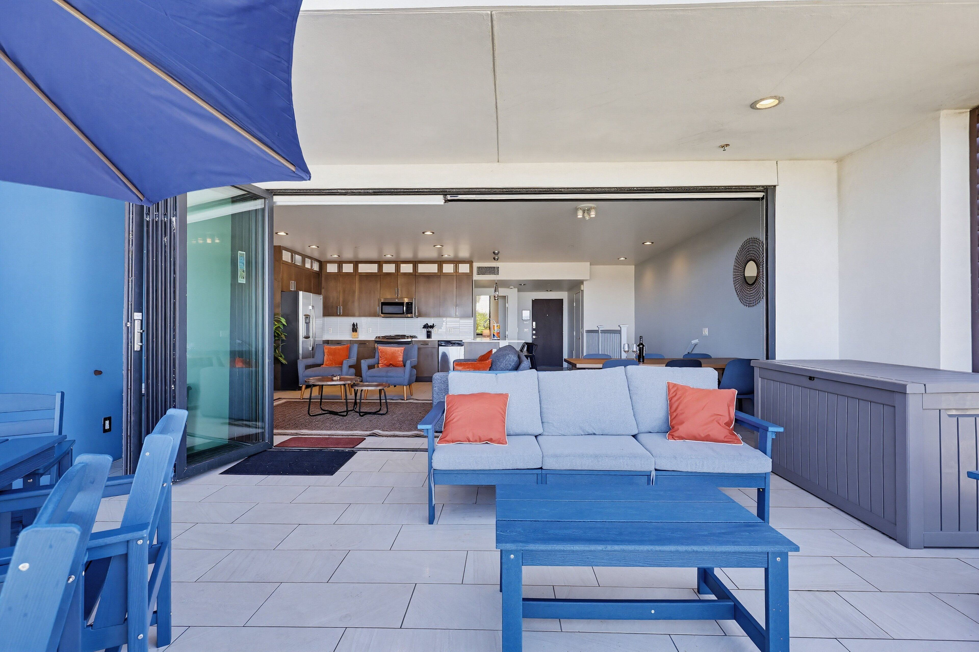 Un espace extérieur aménagé avec un canapé et une table bleus, une cuisine en arrière-plan et une porte-fenêtre coulissante servant d’entrée.