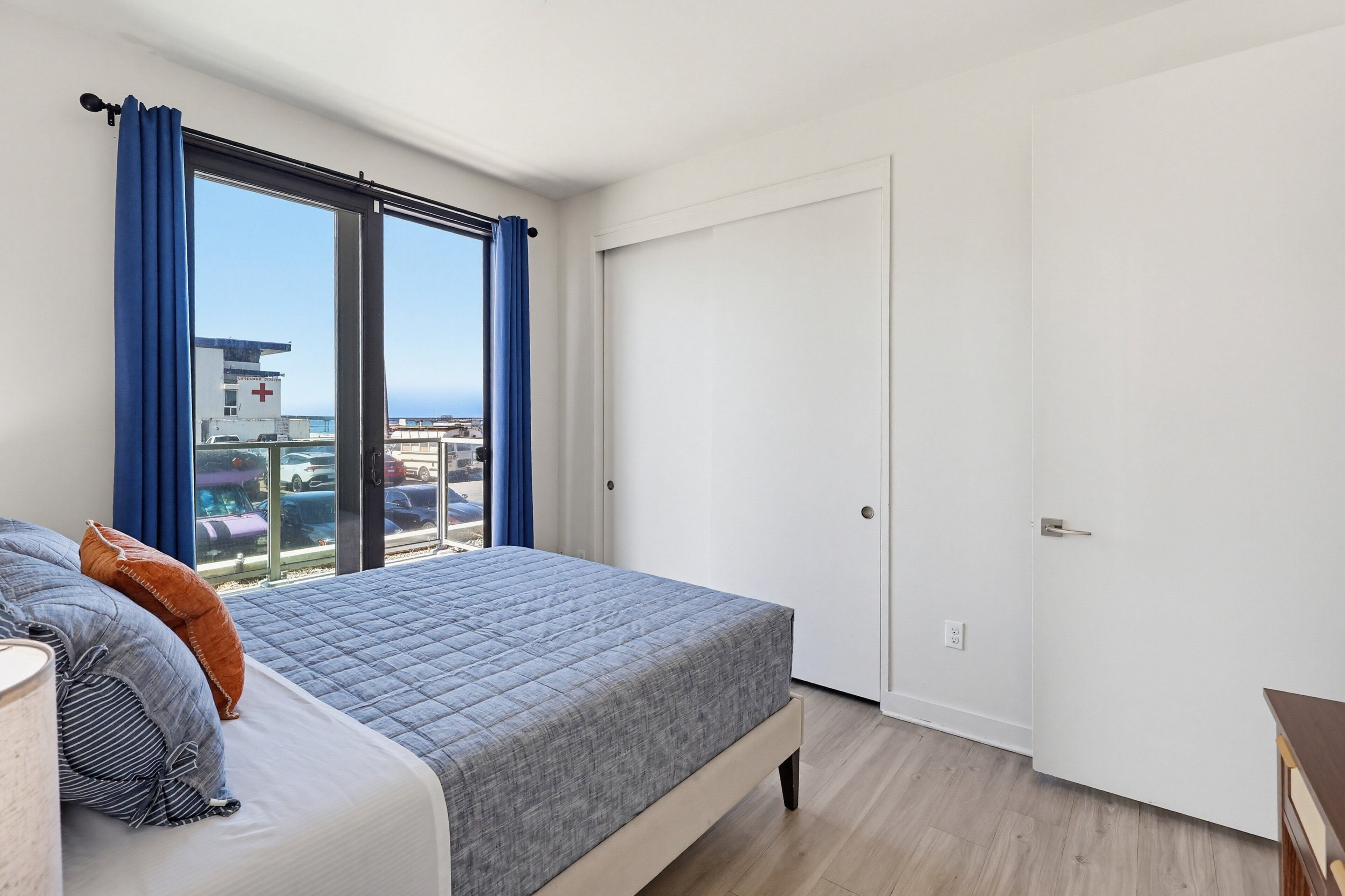 A bedroom with a bed, a window with curtains, a door, and a view of buildings and cars outside.