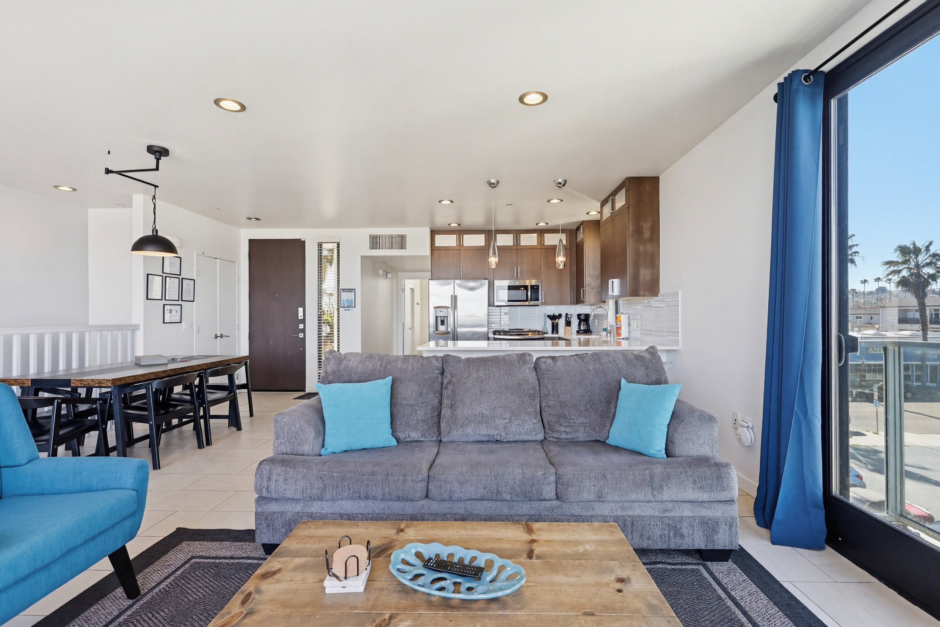 A modern living room with a grey sofa, blue armchairs, a wooden coffee table, and a view of the kitchen and balcony.