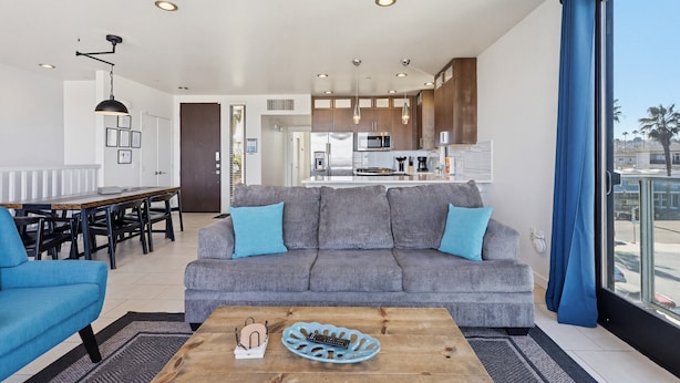 A modern living room with a grey sofa, blue armchairs, a wooden coffee table, and a view of the kitchen and balcony. - Ocean Park Combo 9 & 10 (San Diego)