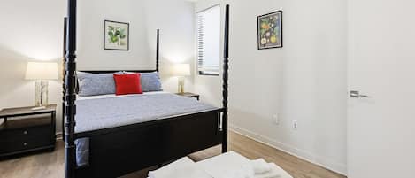 A four-poster bed with a headboard, a bedside table with a lamp, and a wall-mounted picture.
