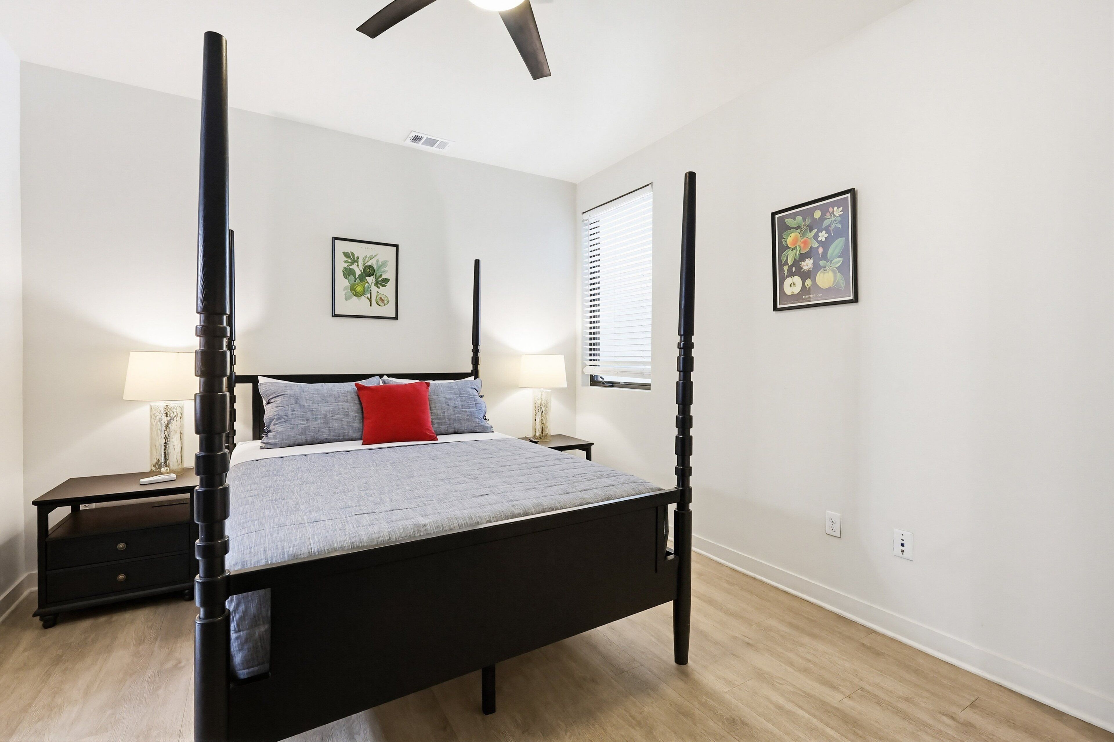 Un lit à baldaquin avec une couvre-lit grise et un coussin rouge, deux tables de chevet avec des lampes, une fenêtre avec des stores et une estampe botanique encadrée au mur.