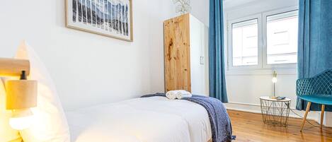 A bedroom with a bed, a wooden wardrobe, a chair, a small table, and a window with curtains.