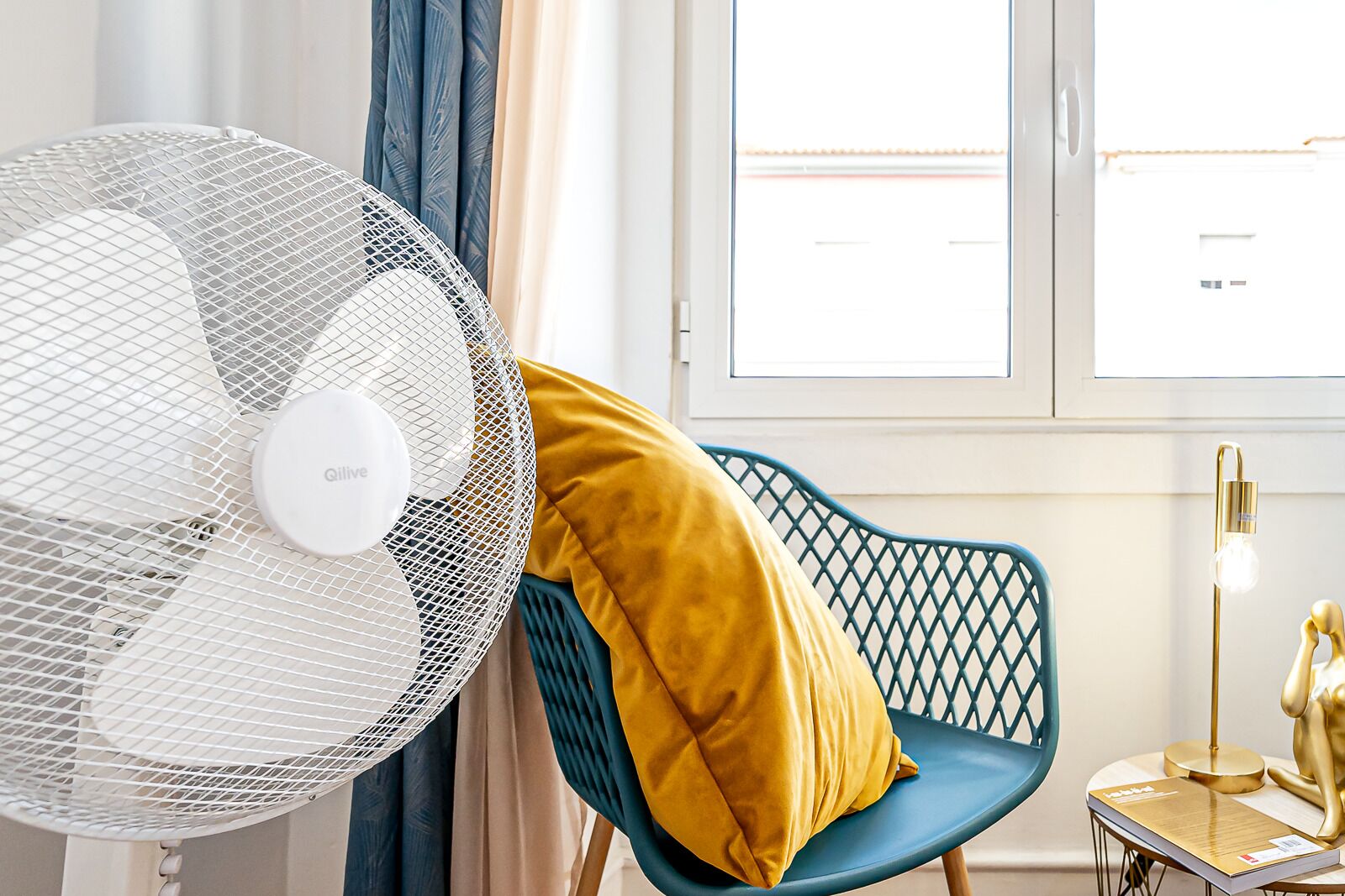 Ein Zimmer mit einem Standventilator, einem blauen Stuhl mit gelber Sitzkissen, einem kleinen Tisch mit einer Lampe und einem Fenster mit Jalousien.