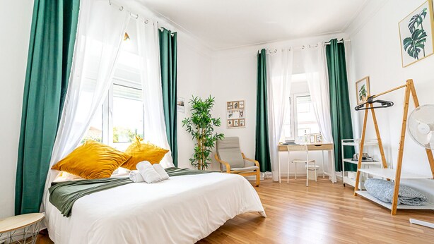 Ein Schlafzimmer mit Bett, Schreibtisch, Stuhl, Pflanze und Ventilator.