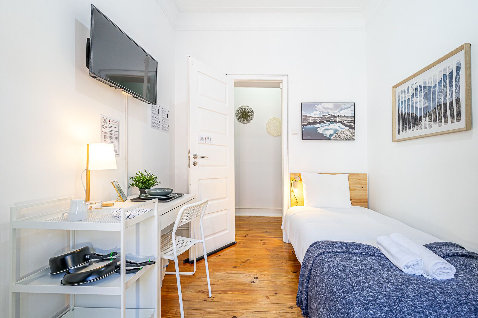 A compact hotel room with a bed, a desk with a chair, a TV mounted on the wall, and a door leading to another room.
