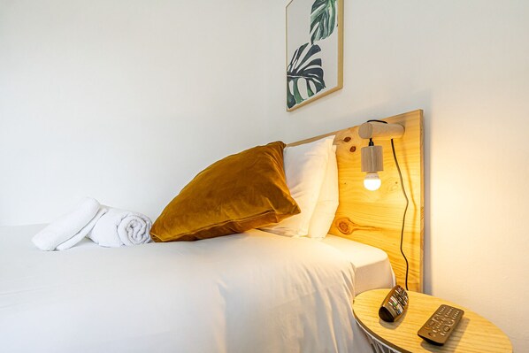 A neatly made bed with white linens, a brown pillow, and a remote control on a wooden side table. - April Square Guesthouse by Lisbon with Sintra (Amadora)