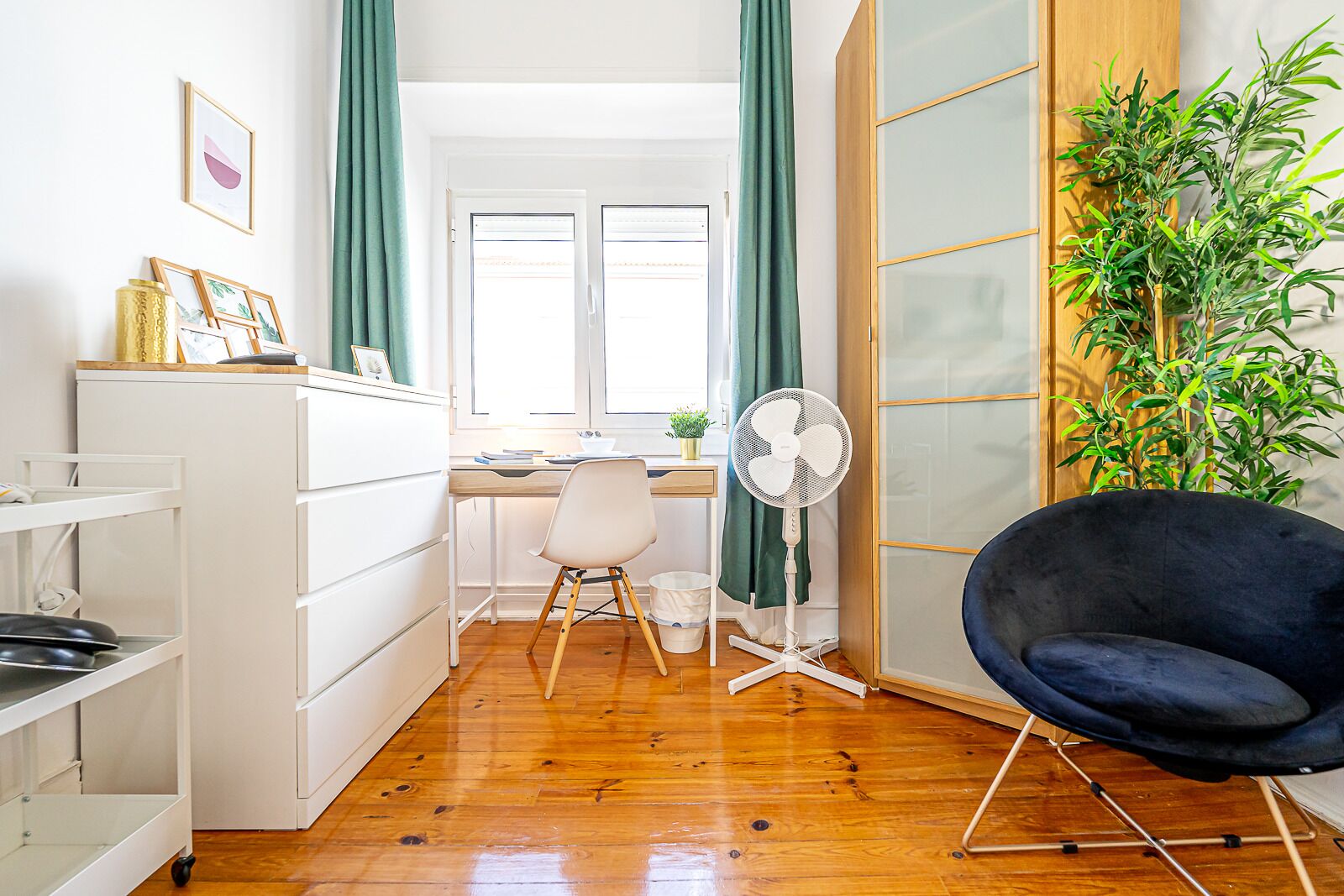 A compact, well-lit room with a wooden floor, a desk with a chair, a standing fan, and a black chair.