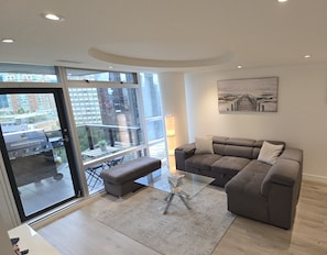 Ein modernes Wohnzimmer mit einem grauen Ecksofa, einem Glastisch und Blick auf eine Stadtlandschaft durch große Fenster.