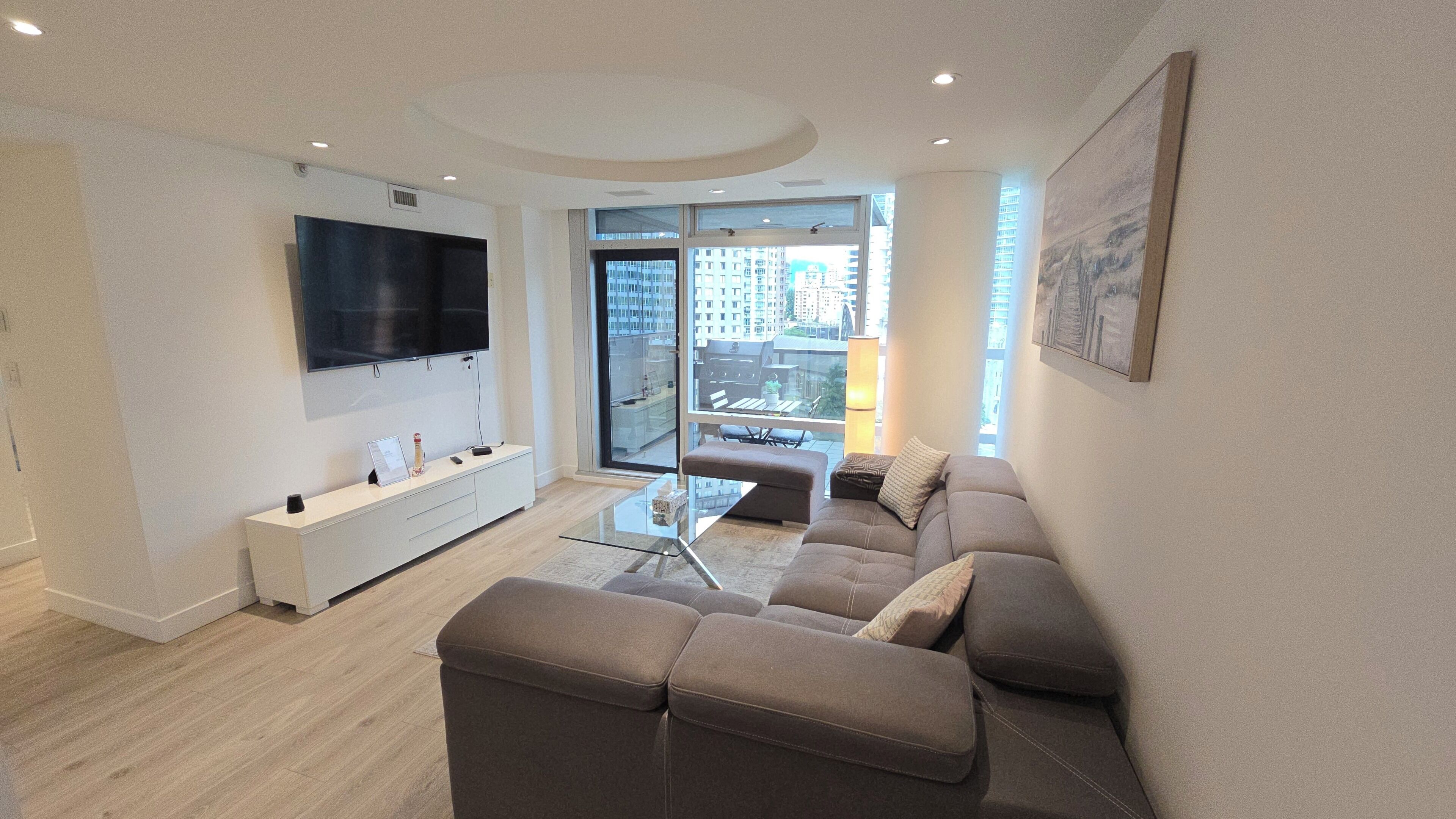 A modern living room with a flat-screen TV, a sofa, a glass coffee table, and a balcony with a view of the city.