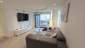 Ein modernes Wohnzimmer mit Flachbildfernseher, einer Couch, einem Glastisch und einem Balkon mit Stadtblick.
