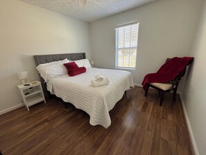 Une chambre à coucher avec un lit, une table de chevet, une chaise, une fenêtre et un ventilateur de plafond.