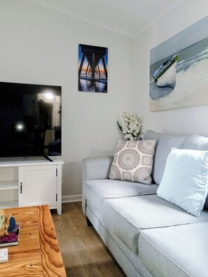 A living room with a grey sofa, a flat-screen TV on a white cabinet, a painting of a bridge, and a vase with white flowers. - Cozy home in the heart of Calabash NC (Calabash)