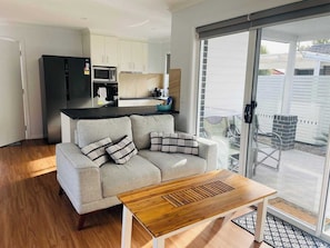 A modern living room with a sofa and a wooden coffee table, a kitchen with white cabinets and appliances, and a sliding glass door leading to a patio.