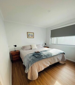 A bedroom with a bed, a nightstand, a lamp, a window with blinds, and a picture on the wall.
