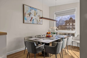 A dining area with a table set for four, a painting on the wall, and a view of mountains through a window. - Luxury Canmore Mountain Retreat|Hot Tub|Sleeps14|4BR+DEN ( BL# RES-11738 ) (Canmore)