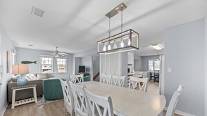 Ein modernes Wohnzimmer mit einer Couch, einem Couchtisch und einem Fernseher. Es gibt einen Essbereich mit Tisch und Stühlen. Der Raum verfügt über einen Deckenventilator und eine Leuchte.