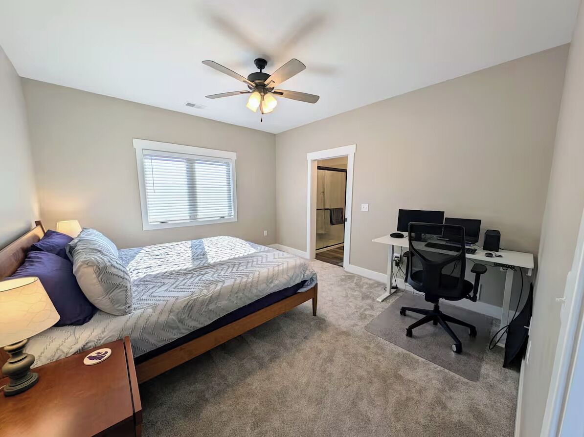 Un dormitorio con cama, escritorio con silla, ventilador de techo y ventana con persianas.