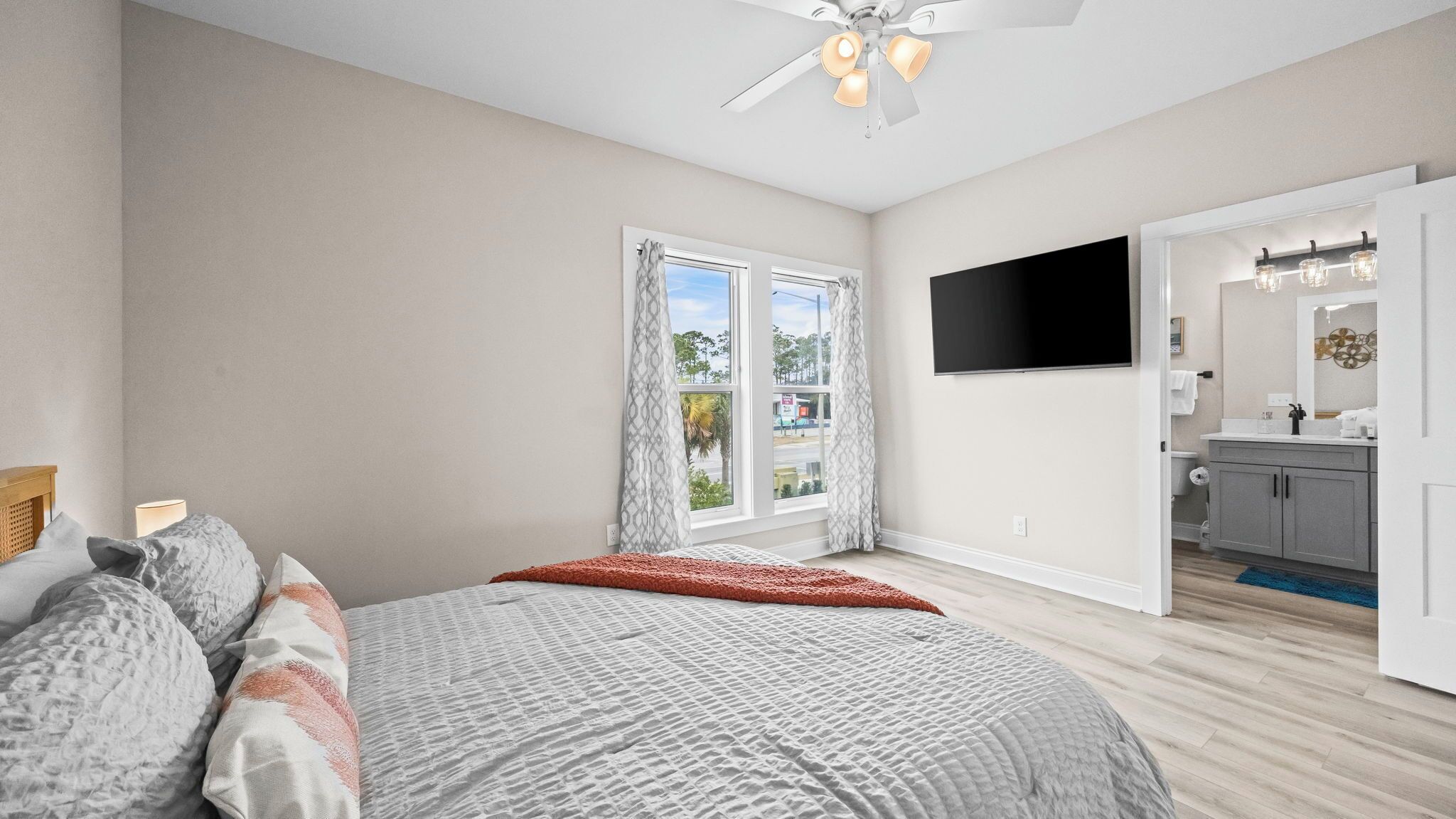 Une chambre à coucher avec un lit, un ventilateur de plafond, une télévision et une salle de bain visible à travers une porte ouverte.