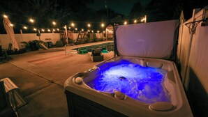 Un bain à remous avec un éclairage bleu, entouré d’une terrasse agrémentée de guirlandes lumineuses, d’un espace salon couvert et d’une piscine.