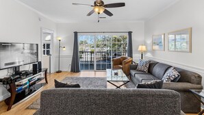 A living room with a flat-screen TV, a ceiling fan, a sofa, a coffee table, and a balcony with a view of greenery. - Magnolia Place 4757-203 (Myrtle Beach)