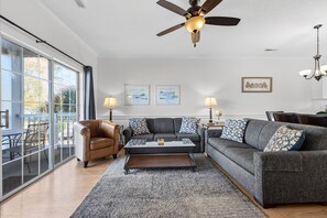 Ein Wohnzimmer mit einer Couch, einem Sessel, einem Couchtisch und einem Deckenventilator. An der Wand hängen zwei gerahmte Bilder und eine Pendelleuchte.