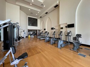 A spacious gym with various cardio machines, weightlifting equipment, and a large window providing natural light. - Burj Khalifa View Luxury 2BR Apartment Souk Al Bahar Downtown Dubai (Dubai)