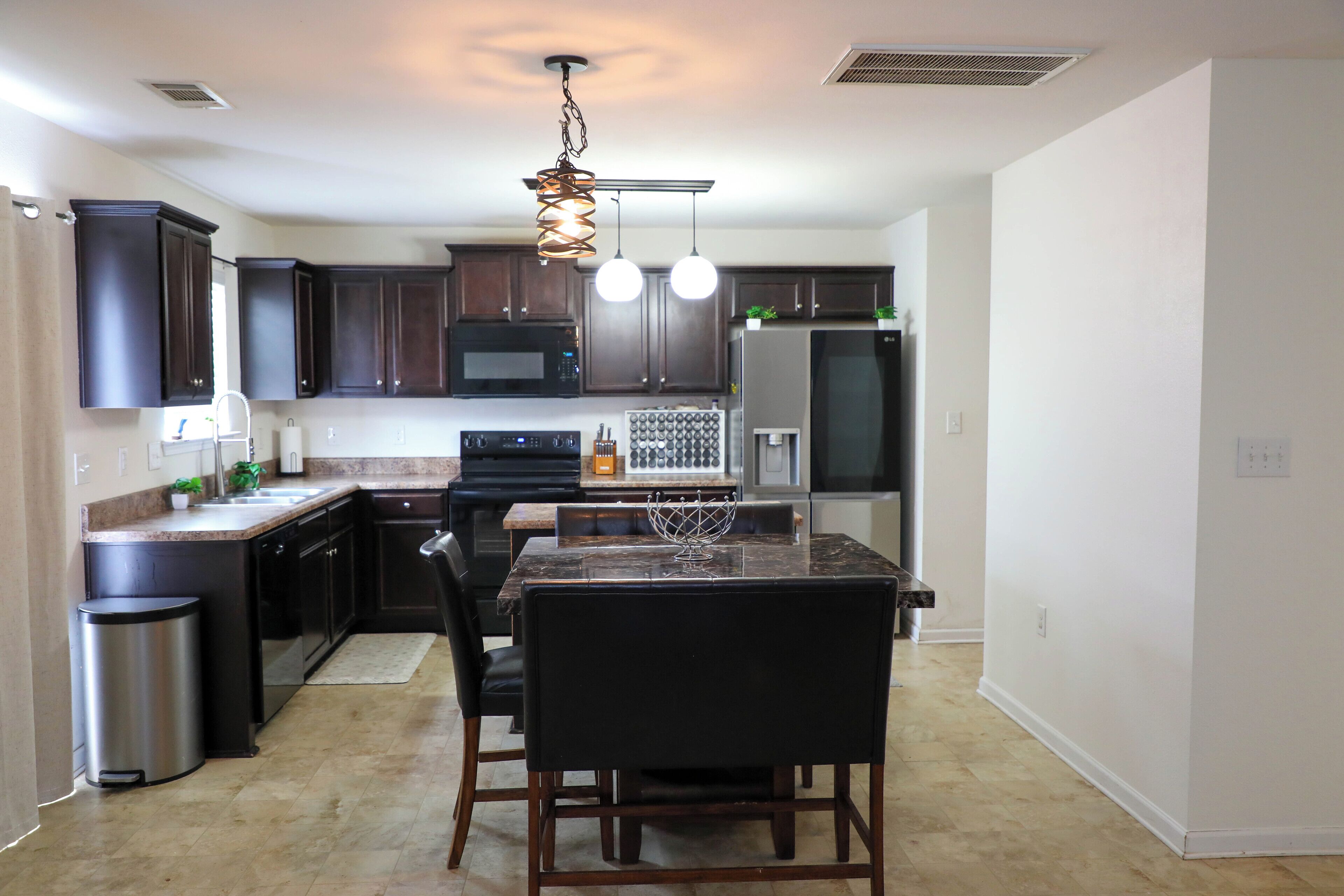 A modern kitchen with dark cabinets, a stainless steel trash can, a dining area with a marble table, and pendant lighting.