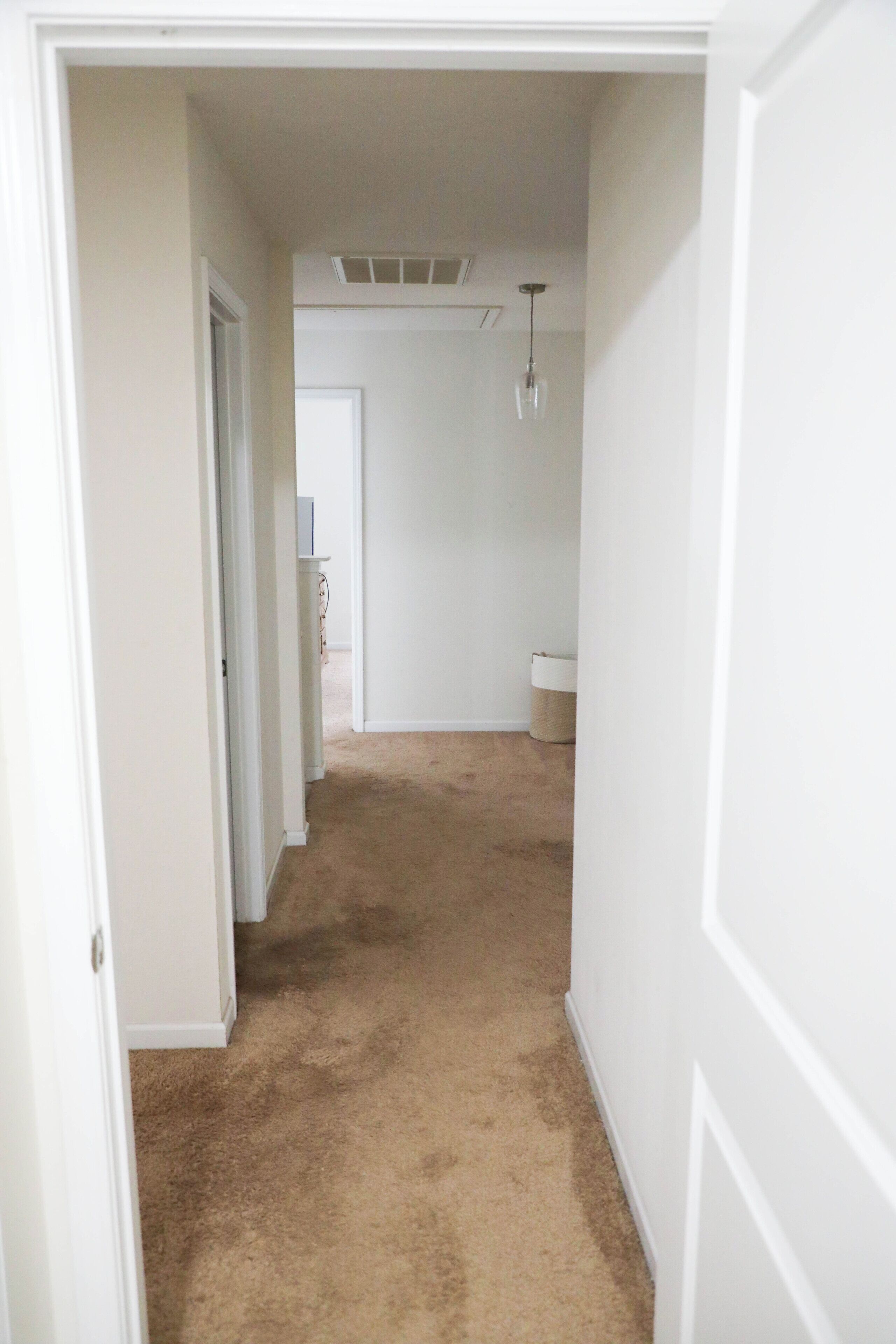 Un pasillo con piso alfombrado, paredes blancas y una puerta que da a otra habitación.