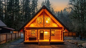 A log cabin with a symmetrical design, a central door, and two windows on the upper level. - Log Cabin! Hot Tub! Firepit! 5 mi Mt Rainier NP! (Ashford)