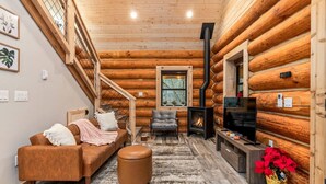 A log cabin interior with a sofa, ottoman, TV, fireplace, and staircase. - Log Cabin! Hot Tub! Firepit! 5 mi Mt Rainier NP! (Ashford)
