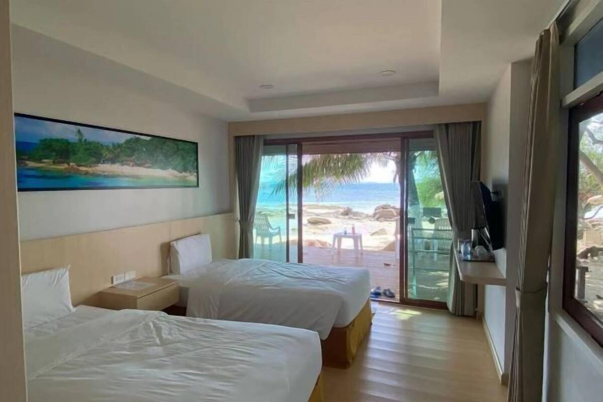 Ein Hotelzimmer mit zwei Betten, einem Strandbild, einem Fernseher und Blick auf den Strand durch eine Glastür.