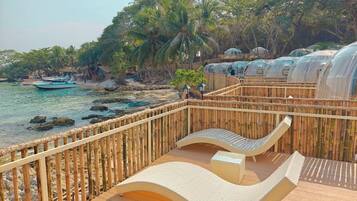 A wooden deck with a curved white lounge chair, a small table, and a bamboo fence overlooking a beach with palm trees and a boat.
