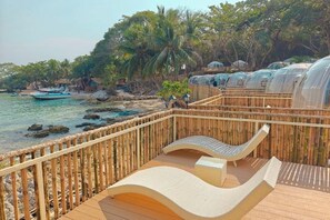 A wooden deck with a curved white lounge chair, a small table, and a bamboo fence overlooking a beach with palm trees and a boat. - Koh Man Klang (Klaeng)