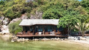 Une cabane en bois, dotée d’un toit de chaume, située au bord d’un lac aux eaux claires, entouré de rochers et d’arbres.